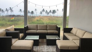 A covered outdoor seating area with wicker furniture and a glass coffee table, overlooking a beach with palm trees and a flag.