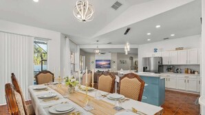 Un comedor moderno con una mesa larga dispuesta para seis personas, muebles de cocina blancos y una zona de cocina con armarios blancos y electrodomésticos de acero inoxidable.