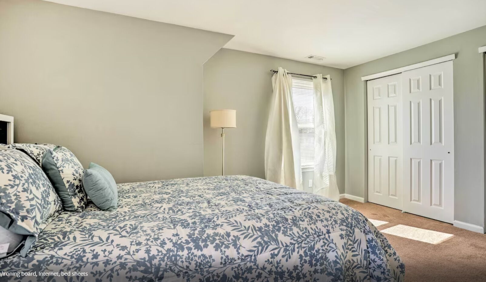 A bedroom with a bed, a nightstand, a lamp, a window with curtains, and a door.