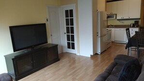 En stue med et fladskærms-tv på et sort skab, en grå sofa og et spiseområde med et bord og stole. Et køkken med hvide skabe og en mikroovn kan ses gennem en åben dør.