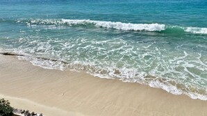Una playa con agua turquesa cristalina, olas suaves y varios veleros anclados en la distancia.