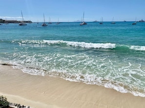 Ein Strand mit klarem türkisblauem Wasser, sanften Wellen und mehreren Segelbooten in der Ferne.