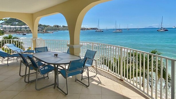 Un espacio de estar techado al aire libre con una mesa y sillas, con vista a un puerto deportivo con barcos y un mar azul y despejado.