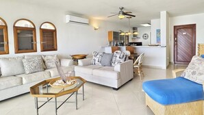 Una sala de estar con un sofá, una mesa de centro y una silla de mimbre. Se observa un área de cocina al fondo, con armarios y una encimera.