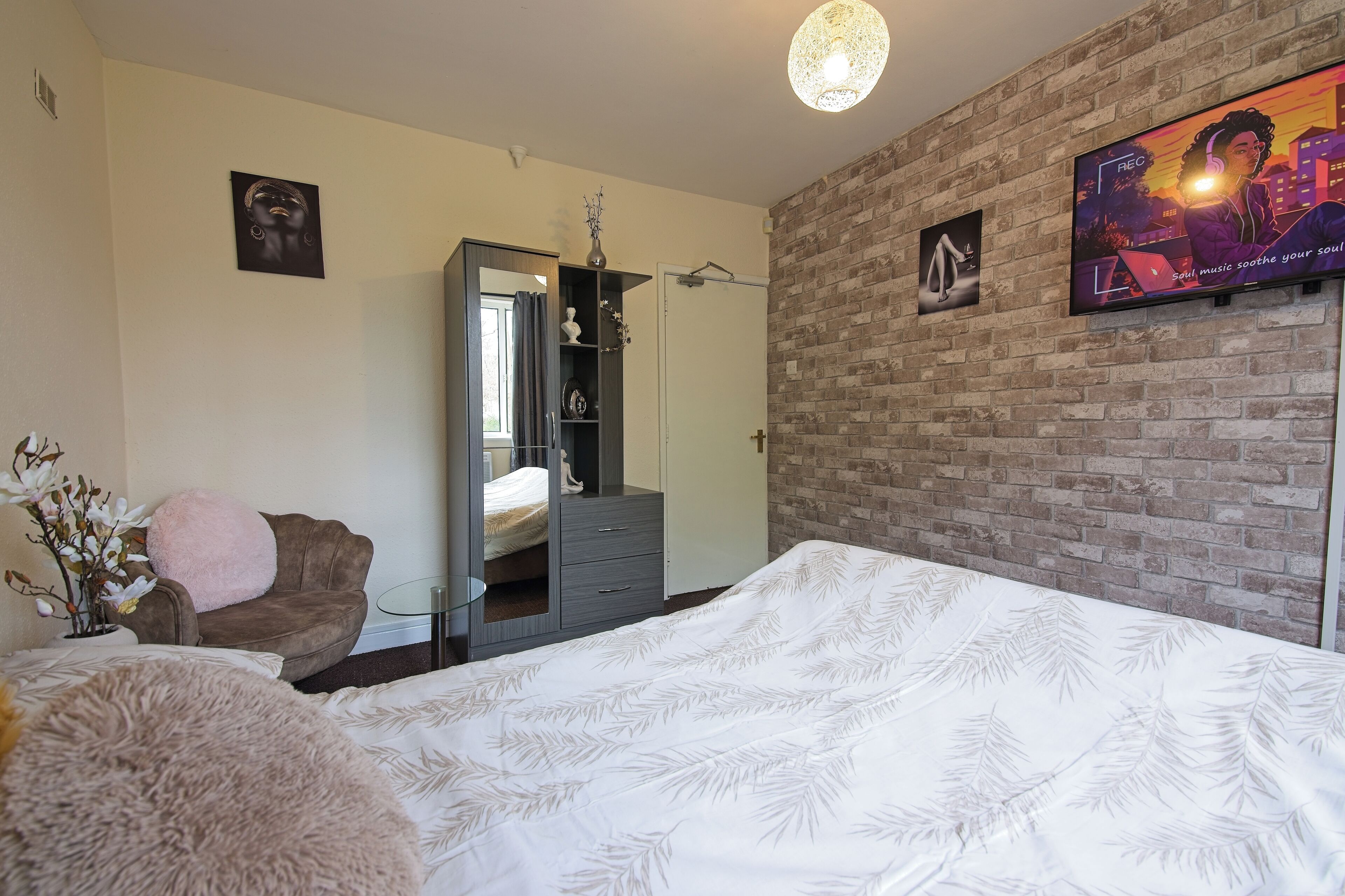 A bedroom with a large bed, a wardrobe, a mirror, a TV on the wall, and a brick wall.