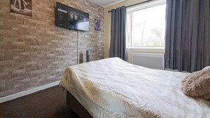 A hotel room with a brick wall, a flat-screen TV, a large window with curtains, and a bed with a leaf-patterned bedspread. - Stylish Five Star Rated Apartment (Manchester)