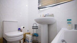 A bathroom with a toilet, sink, and bathtub. There are toilet paper and cleaning supplies visible. - Stylish Five Star Rated Apartment (Manchester)