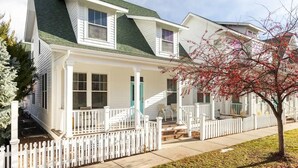 A two-story house with a porch, white picket fence, and a tree with red leaves.