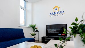En moderne stue med en blå sofa, et spisebord med mad på, et fjernsyn på et stativ, og et vægmonteret logo for 'AMOUR NIGHTS'.