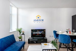 A modern living room with a flat-screen TV, a sofa, a dining table with chairs, and a logo on the wall.