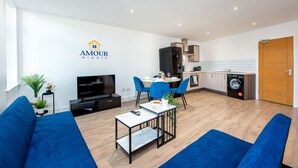 En moderne stue med TV, blå sofaer, et spisebord med stole, et køkken med hårde hvidevarer, og et logo for 'AMOUR NIGHTS'.