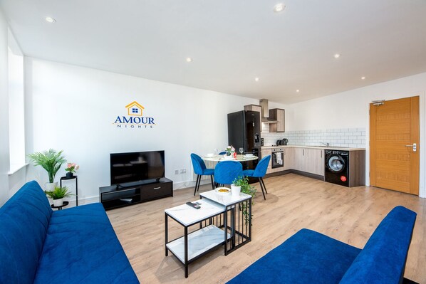 A modern living room with a TV, blue sofas, a dining table with chairs, a kitchen area with appliances, and a logo for 'AMOUR NIGHTS'.