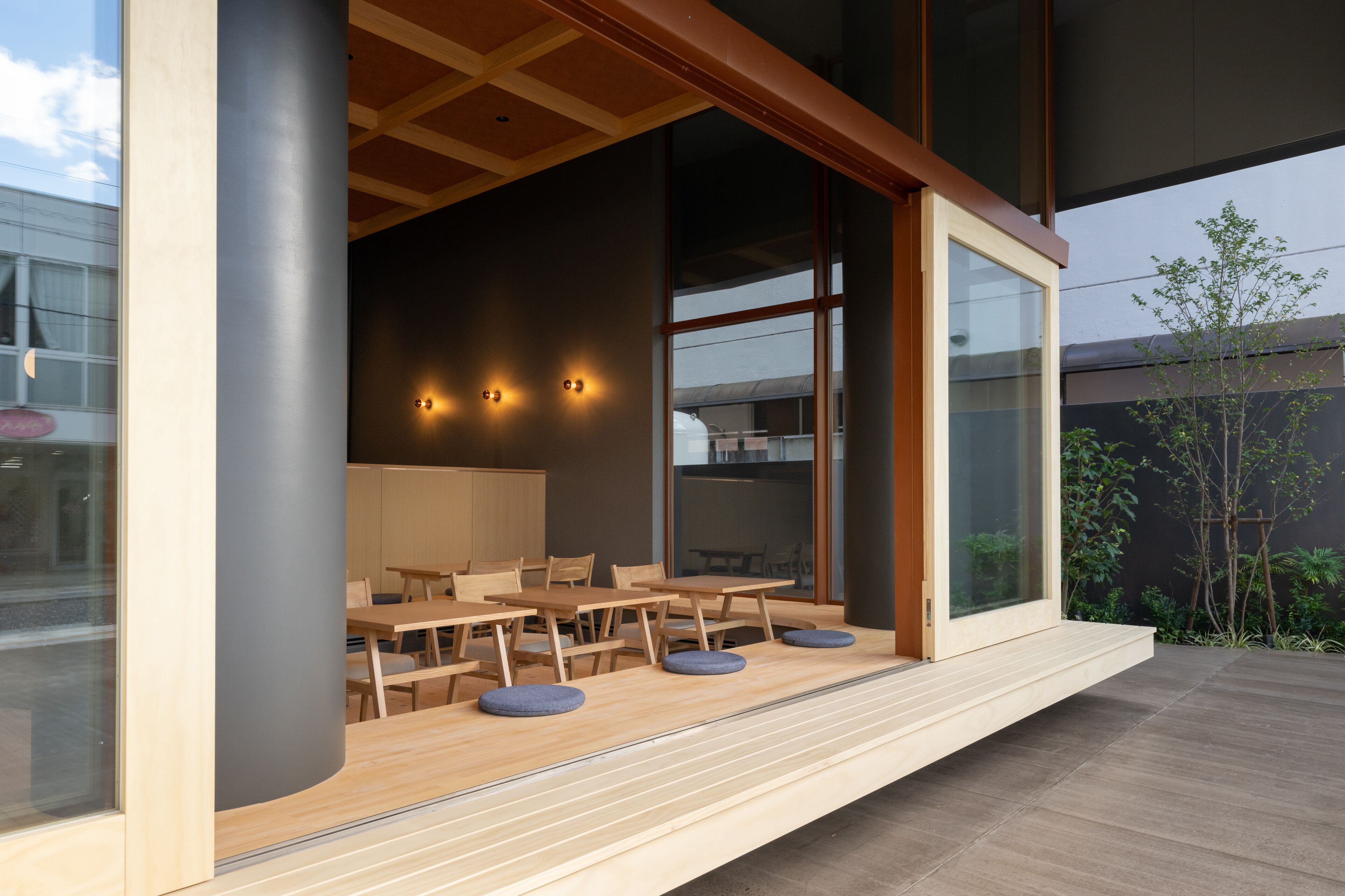 An outdoor dining area with wooden tables and chairs, a glass wall, and a view of the building's interior.