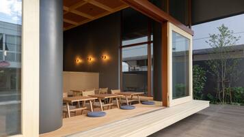 An outdoor dining area with wooden tables and chairs, a glass wall, and a view of the building's interior.