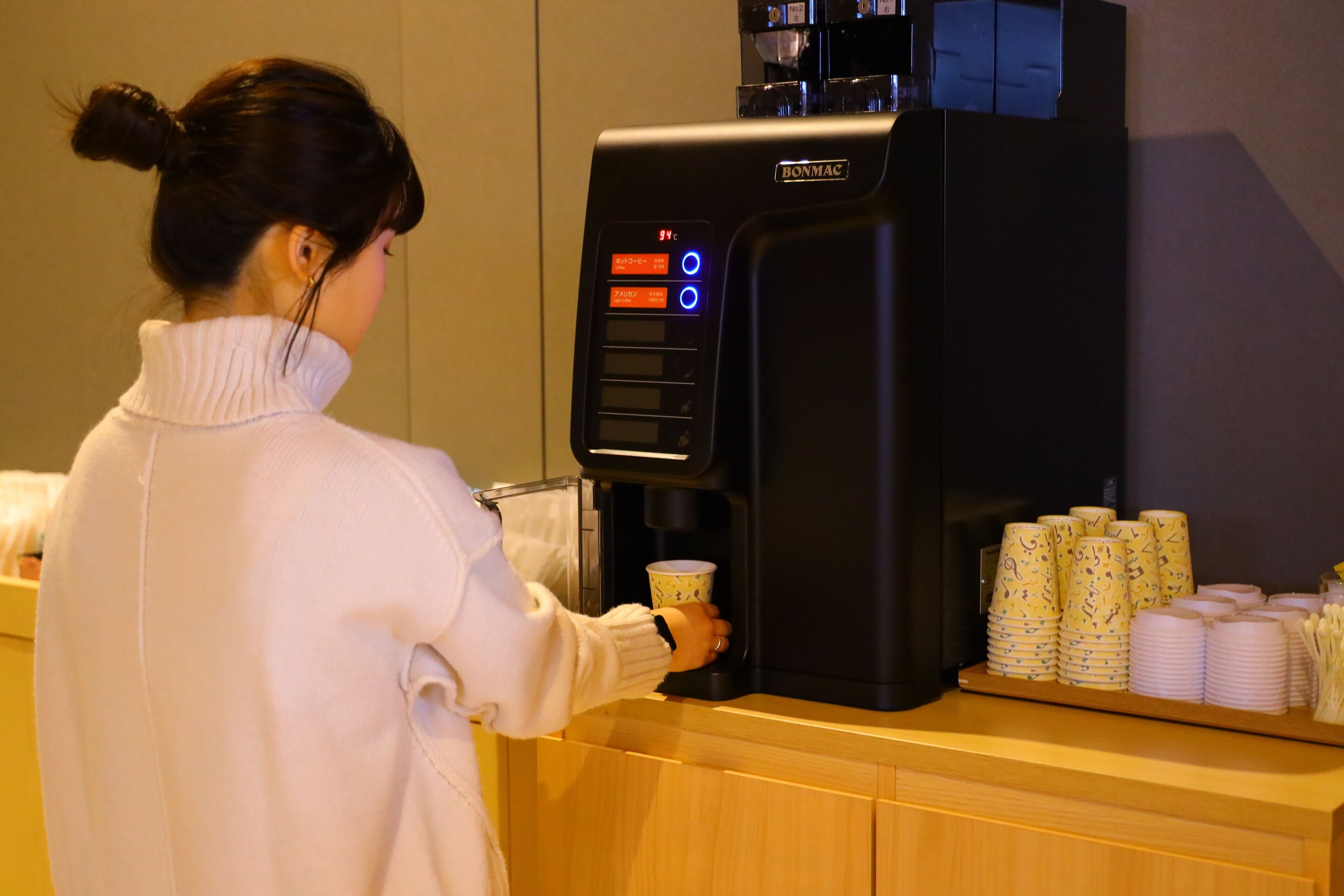A person using a coffee machine with a digital display, a coffee cup, and stacks of disposable cups and plates on a counter.