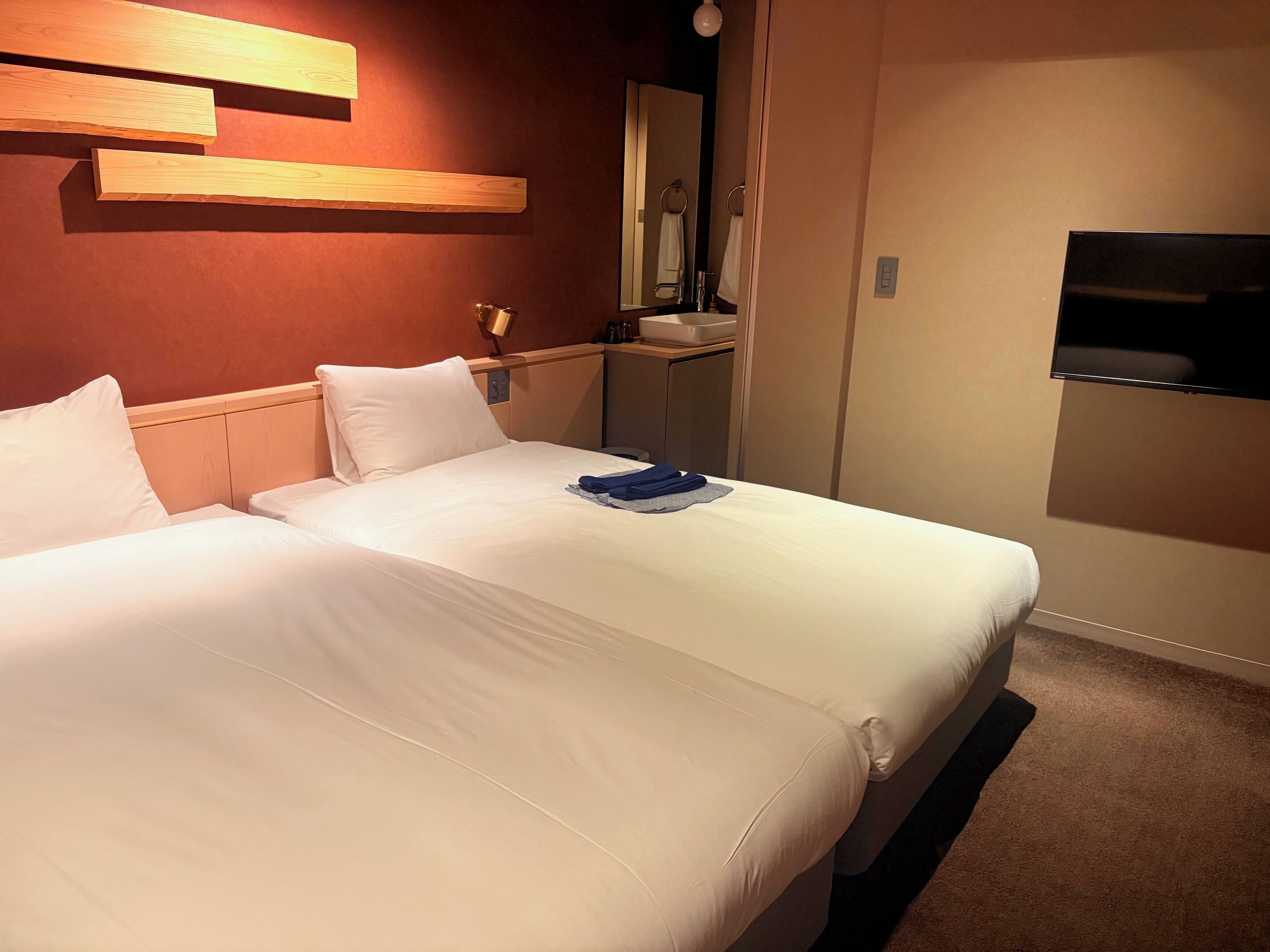A hotel room with two beds, a wooden headboard, a mirror, a TV, and a small shelf with toiletries.