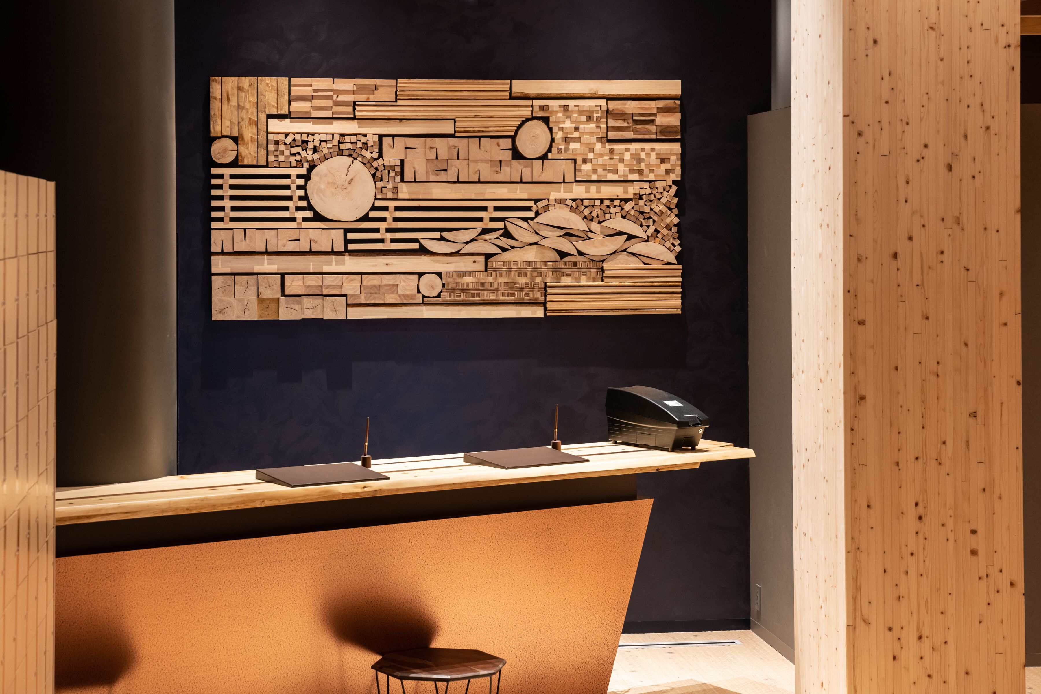 A reception desk with a wooden counter, a dark wall with a geometric wood art piece, and a single chair.