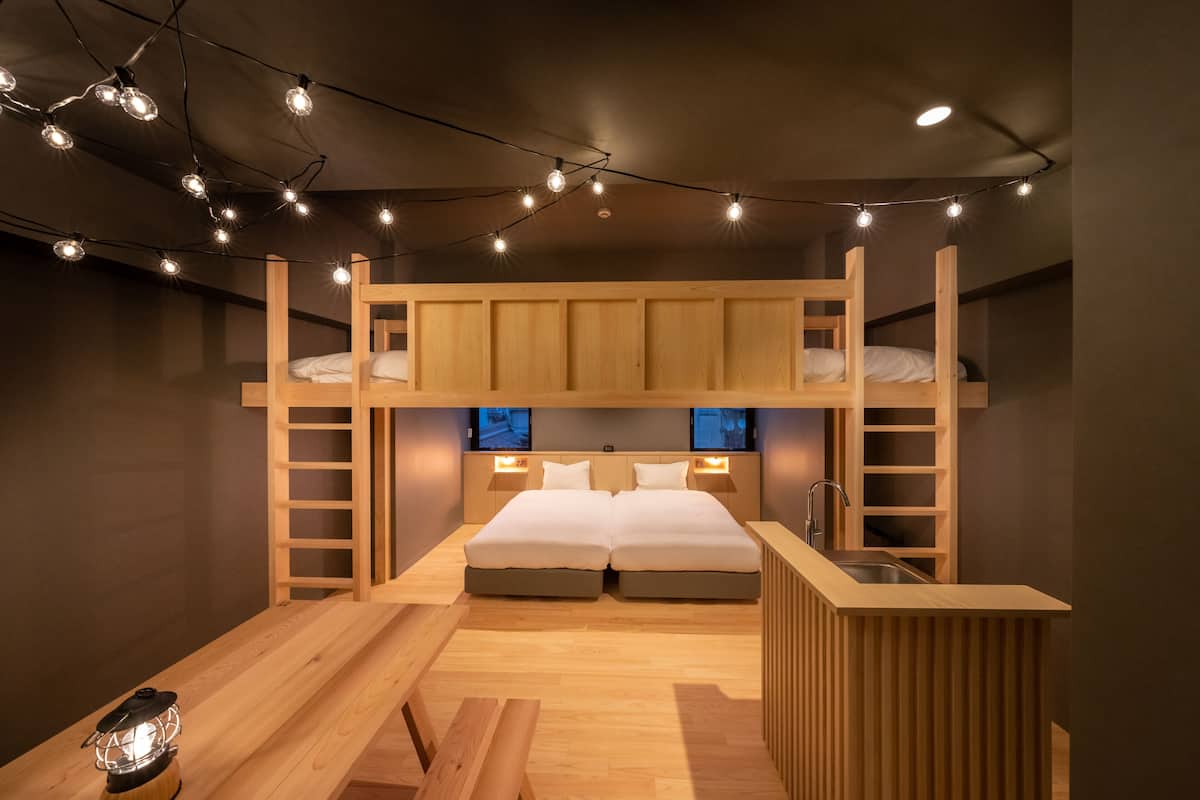 Habitación con literas, un escritorio de madera y un banco, luces colgantes y una lámpara de mesa pequeña.