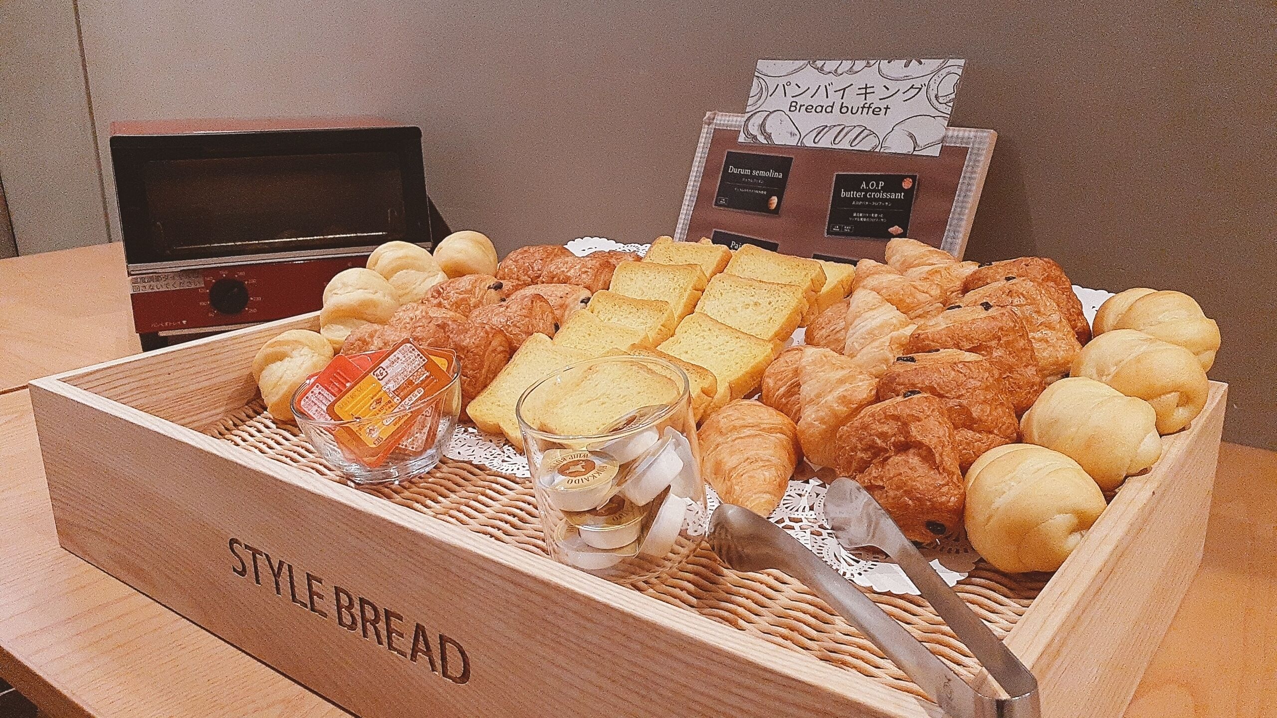 Une boîte en bois étiquetée « PAINS DE STYLE » contenant divers pains et pâtisseries, un verre contenant une boisson et un panneau indiquant un « buffet de pains ».