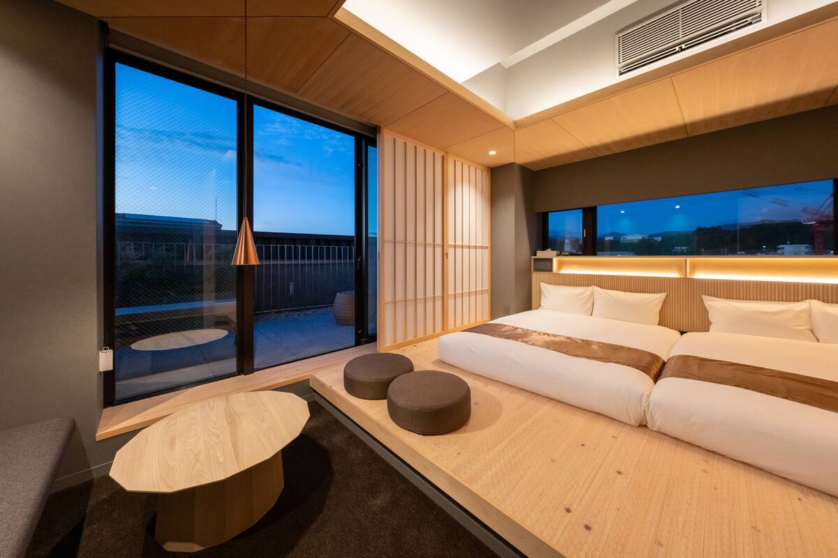 Una habitación de hotel moderna con un amplio ventanal corredero que da a un balcón, un sofá cama con almohadas, una pequeña mesa redonda y un televisor montado en la pared.