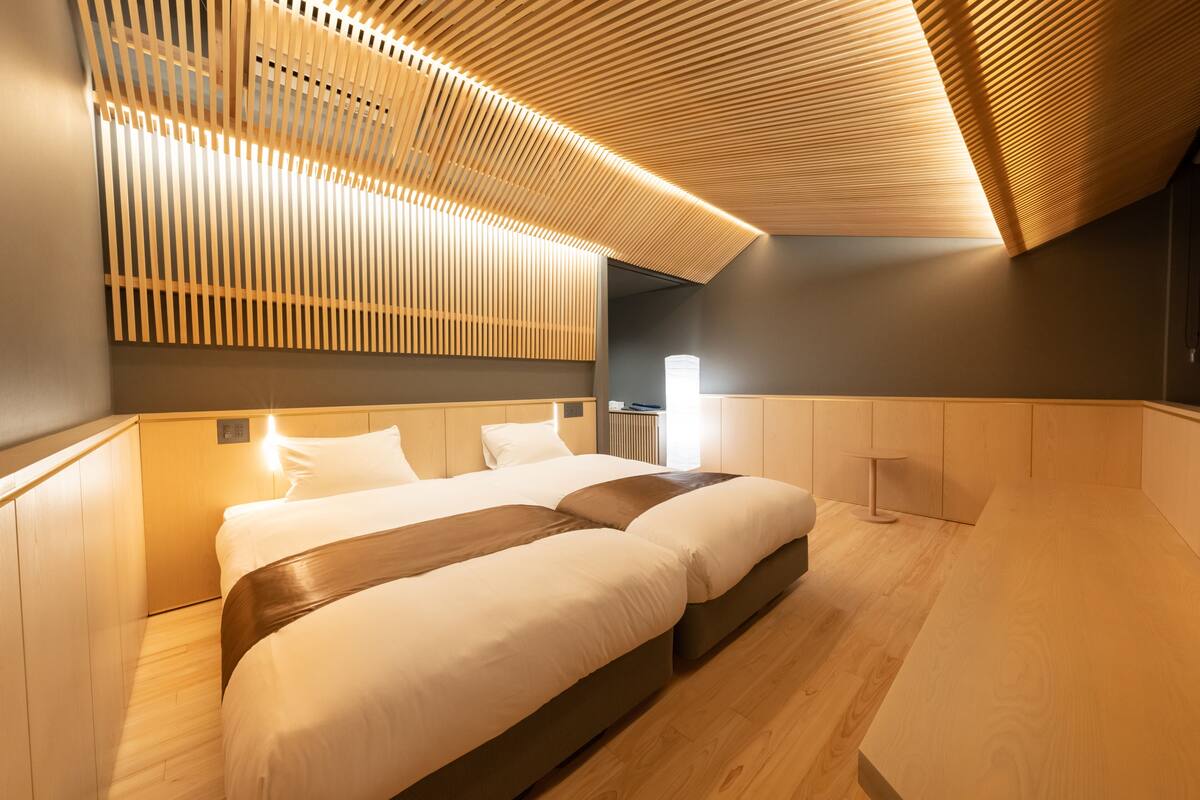 Una habitación de hotel moderna con techo de madera, dos camas, una mesita de noche y un buró.