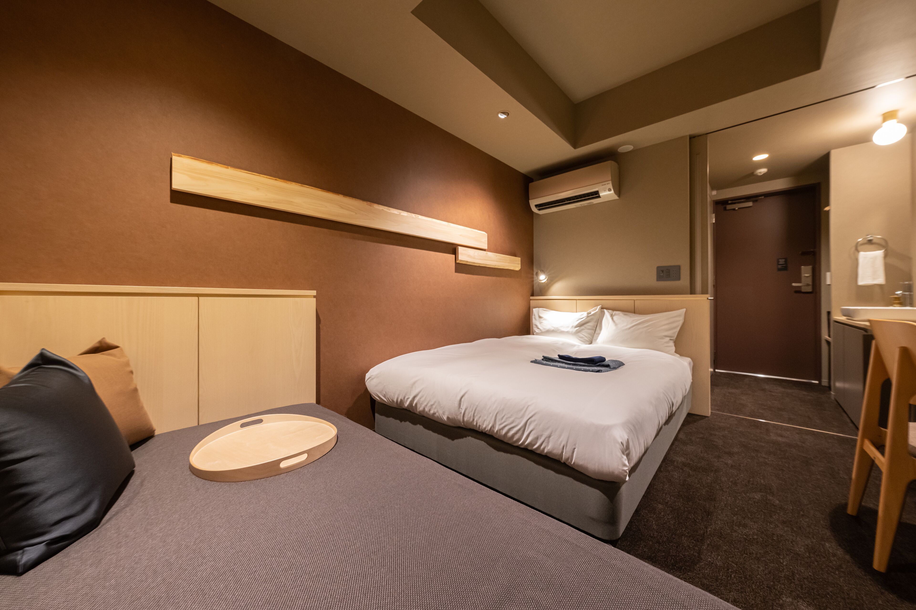 A hotel room with a bed, a bedside table, a chair, a wooden shelf, and an air conditioning unit.