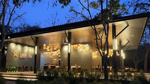 A modern building with an open patio area featuring multiple dining tables and chairs, surrounded by lush greenery.