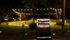 A nighttime view of a campsite with a sign reading "THE CAMPFIRE," surrounded by string lights and trees.