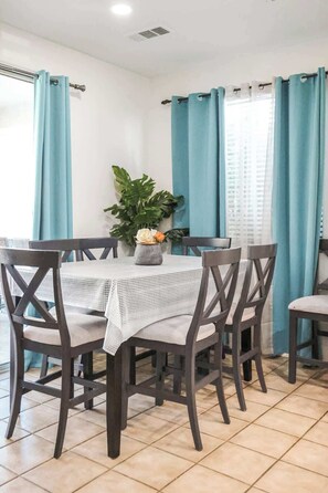 Een eetruimte met een tafel gedekt voor vier personen, omringd door donkerhouten stoelen, een geweven tafelkleed en een potplant met witte bloemen.