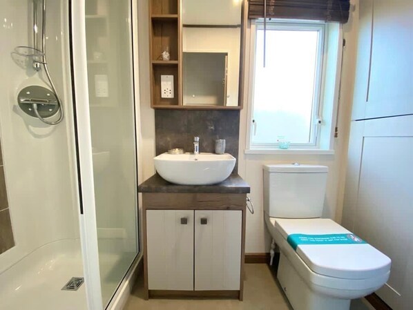 A compact bathroom with a glass-enclosed shower, a white sink with a marble countertop, a toilet, and a window providing natural light. - Pet-Friendly 3-Bed Lodge for 8, Tattershall Lakes (Lincoln)