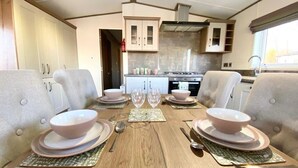 Un coin repas avec une table en bois dressée pour quatre personnes, entourée de chaises rembourrées. On aperçoit, au fond, une cuisine avec des placards et une cuisinière.