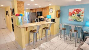 A kitchen with a central island, bar stools, wooden cabinets, a microwave, and a clock on the wall. - Habitat #23 1 Bedroom Home by RedAwning (South Padre Island)