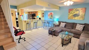 A living room with a sofa, coffee table, and chairs. There is a kitchen area in the background with a bar and stools. - Habitat #23 1 Bedroom Home by RedAwning (South Padre Island)