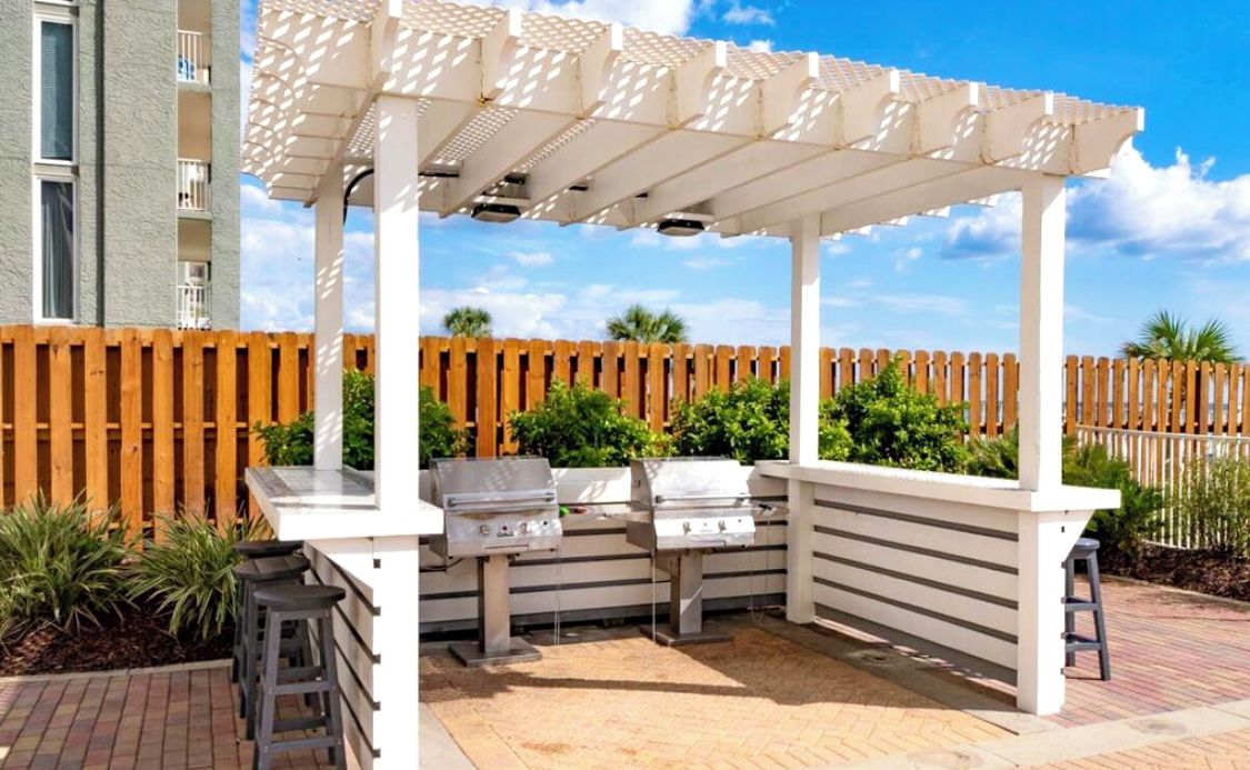 An outdoor patio with a pergola, two barbecue grills, and a wooden fence in the background.