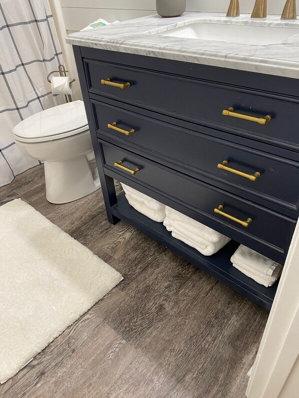 Un baño con tocador azul oscuro, encimera de mármol blanco y tiradores dorados. Hay un inodoro blanco y una alfombra blanca en el suelo.
