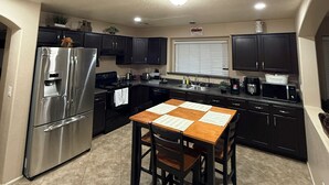 A modern kitchen with dark cabinets, stainless steel appliances, a wooden dining table, and a window with blinds. - Cozi 3 bedroom with pool table and full bar (Maricopa)