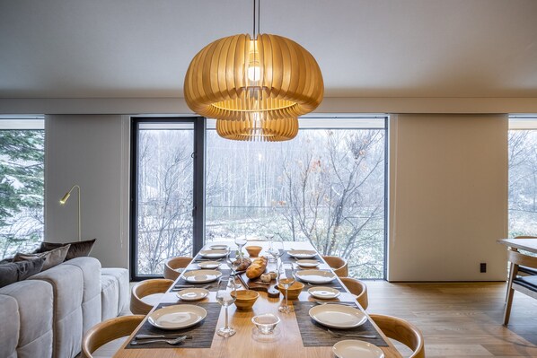 A dining area with a long table set for a meal, large windows, and a modern pendant light.