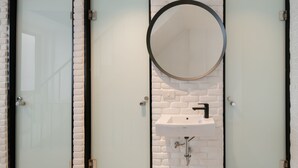 A restroom with three glass doors, a white brick wall, a round mirror, and a sink. - CASA BLANCA Dusit - Hostel (Bangkok)