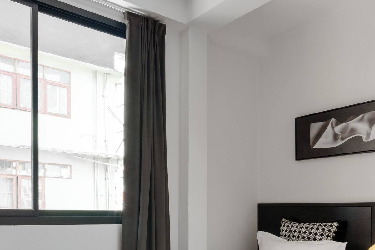Quarto de hotel moderno com duas camas, uma janela grande com cortinas, um ventilador de teto e uma obra de arte emoldurada na parede.