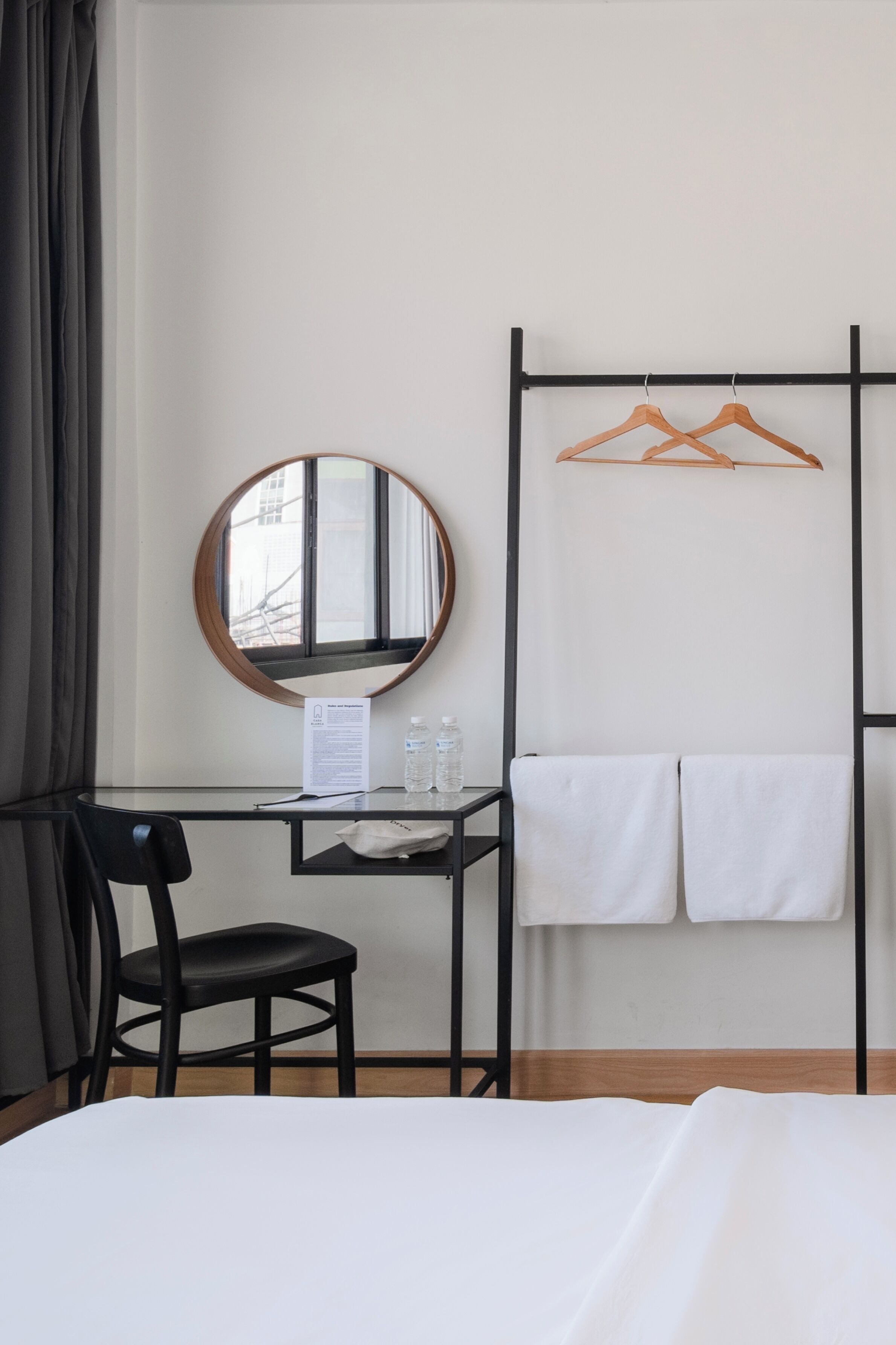 Hotellihuone, jossa on sänky, työpöytä tuolilla, pyöreä peili, musta metallinen vaateteline ja ikkuna, jossa on verhot.