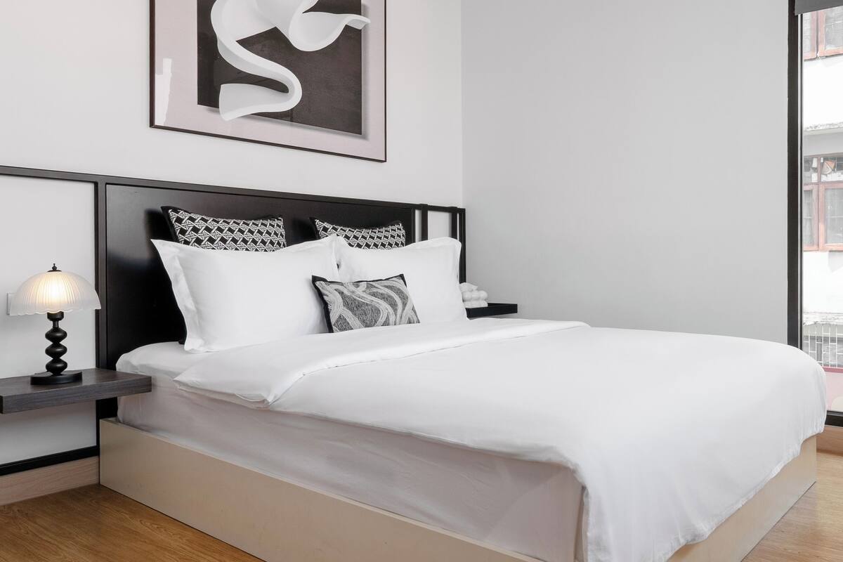 Quarto moderno com ventilador de teto de madeira, obra de arte abstrata emoldurada, cama com roupa de cama branca, almofadas estampadas e mesa de cabeceira com luminária.