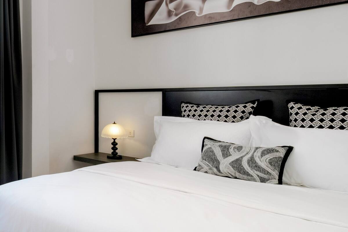 Quarto de hotel bem organizado, com uma cama, mesa de cabeceira com abajur, ventoinha no teto e obra de arte abstrata na parede.