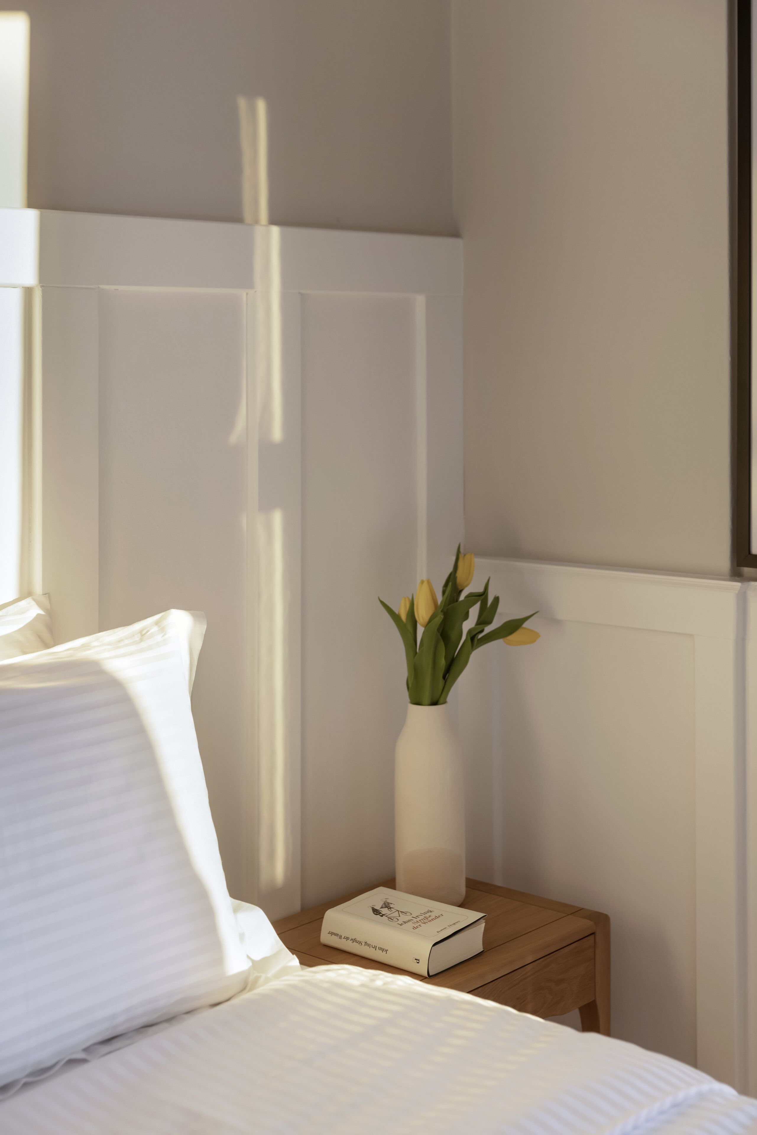 A neatly made bed with white linens, a bedside table with a white vase containing yellow flowers, and a book on the table.