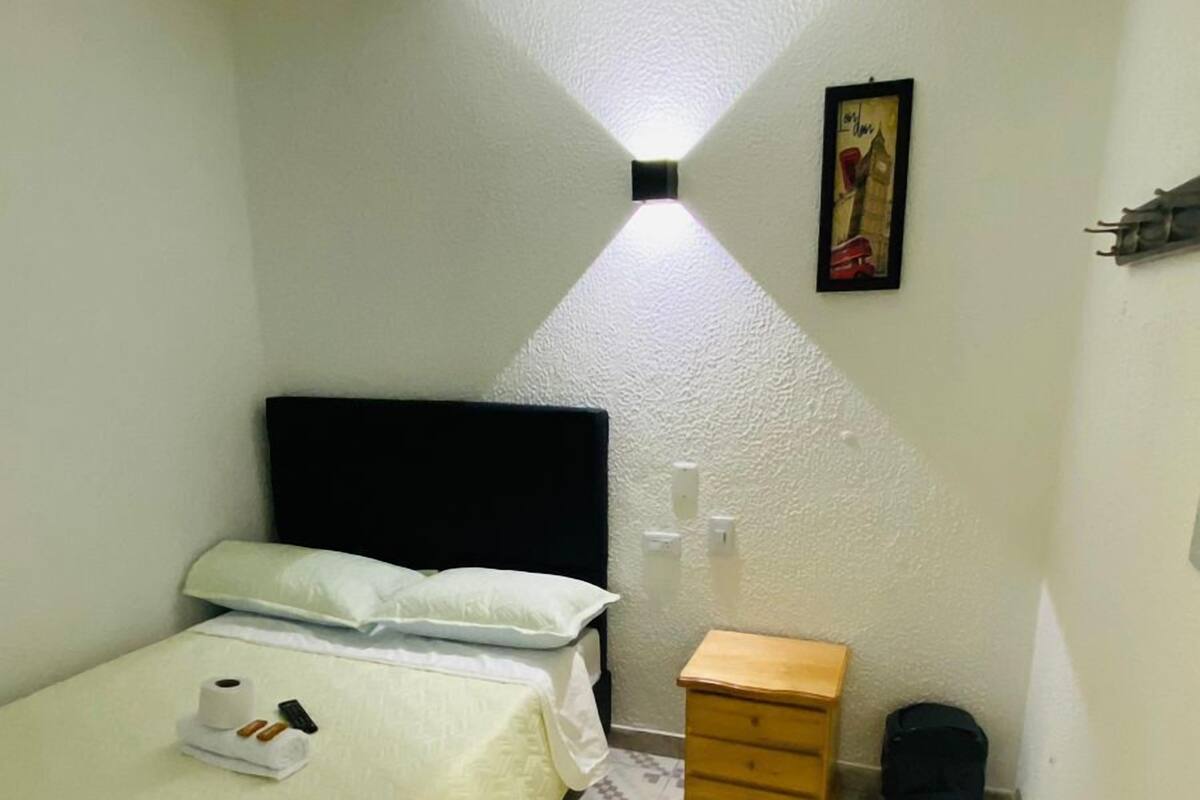 Une petite pièce étroite, avec un lit, une table de chevet, une petite armoire en bois et un sol carrelé.