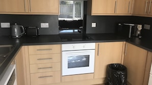 A kitchen with wooden cabinets, a black countertop, a built-in oven, a coffee maker, and a trash bin. - Lovely 1-bed Cottage in Hopeman (Elgin)