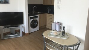 A compact living space with a flat-screen TV, a washing machine, a small dining table with a bottle and a plate, and a kitchen area with cabinets and a kettle. - Lovely 1-bed Cottage in Hopeman (Elgin)