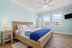 Quarto com cama, mesa de cabeceira, luminária, ventoinha no teto e televisão fixada na parede.