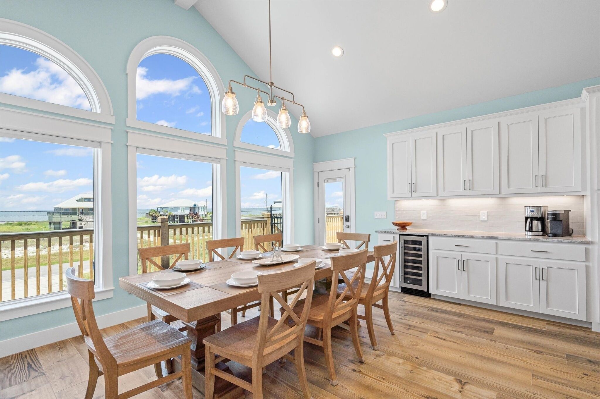 A dining area with a table set for four, wooden chairs, and a view of the beach through large windows.