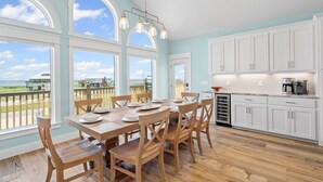 A dining area with a table set for four, wooden chairs, and a view of the beach through large windows. - Surfside - 2431 Bienville 4 Bedroom Home by RedAwning (Dauphin Island)