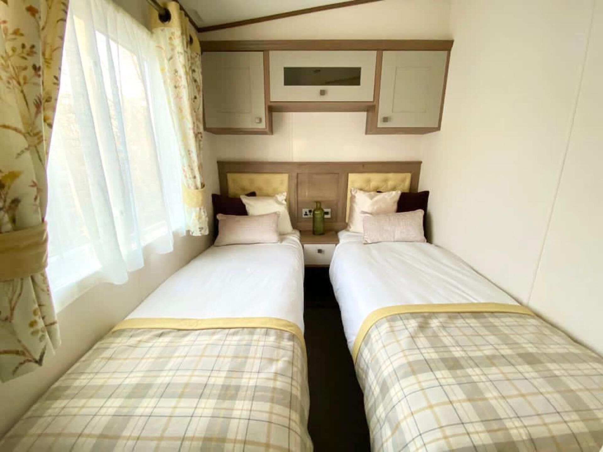 A compact hotel room with two beds, a small bedside table with a lamp, a window with curtains, and a built-in TV above the beds.